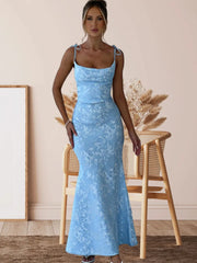 Woman wearing a blue lace dress standing in a room with a wooden chair and framed picture.