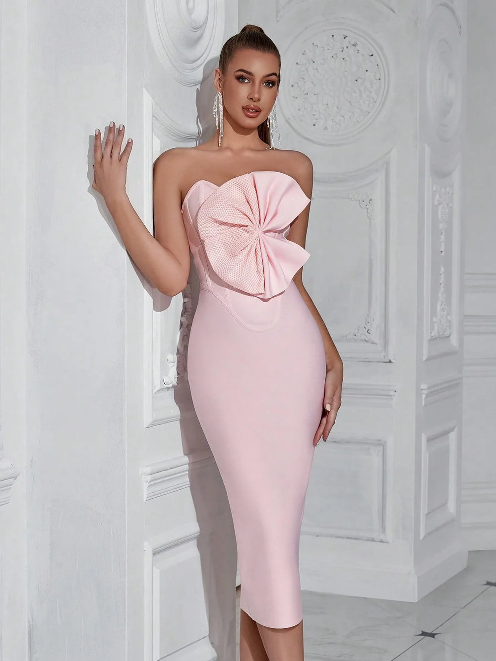 Woman wearing a pink strapless dress with a large bow detail against a white wall.