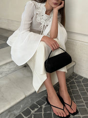Woman in a white outfit with black sandals and a black handbag sitting on a stone floor.