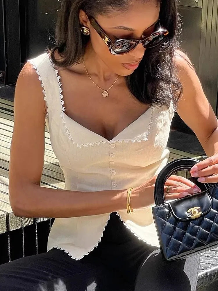 Woman in a white sleeveless top and sunglasses holding a black handbag with a visible brand logo.