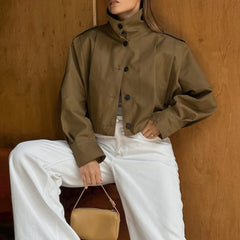 Person wearing a brown jacket and white pants, sitting against a wooden wall.