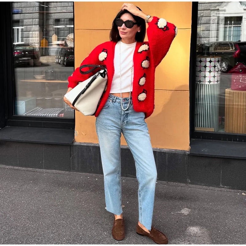 Woman wearing a red cardigan with white flowers, white shirt, blue jeans, and brown shoes, standing against a building exterior.