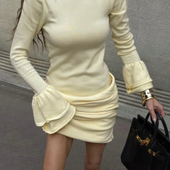 Person wearing a light-colored long-sleeve top and skirt holding a black handbag against a neutral background.