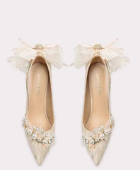 Pair of beige high-heeled shoes with decorative bows and pearls on a white background