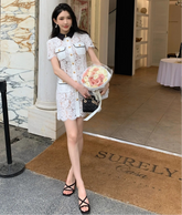 Woman in a white lace dress holding flowers outside a building with 'SURELY Casa' doormat.