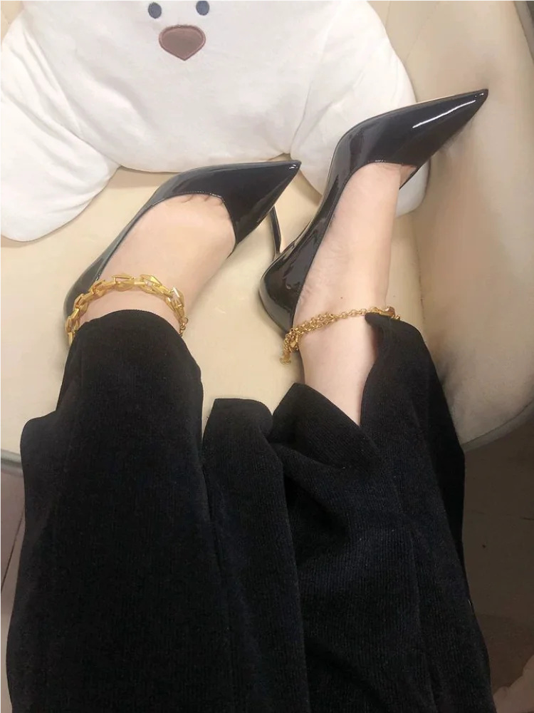 Person wearing black high-heeled shoes and gold ankle bracelets on a light-colored surface.