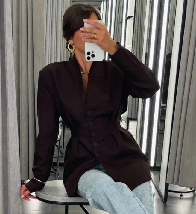 Person wearing a dark brown coat and light blue jeans sitting on a stool in a room with curtains.