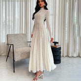 Woman in a long, light-colored lace dress standing in a room with a chair and curtain.