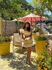 Woman in a yellow outfit standing in a garden with flowers and outdoor furniture.