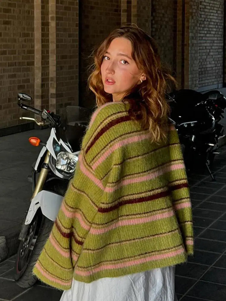 Woman wearing a green and pink striped sweater standing next to a motorcycle.