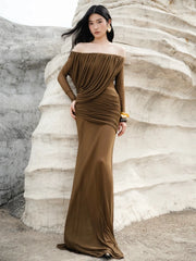 Woman in a brown off-shoulder dress standing against a textured white wall.