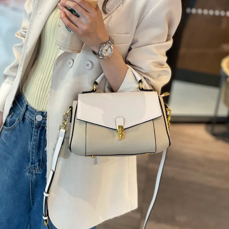 Person holding a white handbag with gold accents, wearing a beige coat and blue jeans.