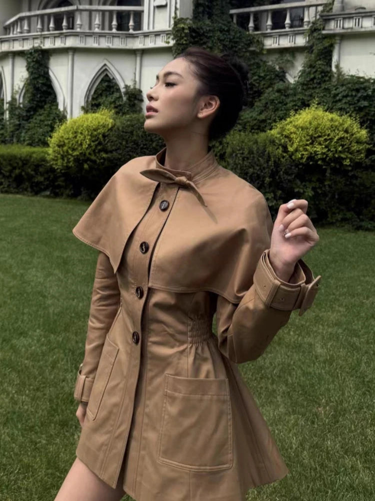 Woman wearing a brown coat standing in a garden with a building in the background