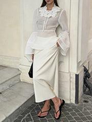 Person wearing a white outfit with a sheer top and black sandals, standing against a light-colored wall.