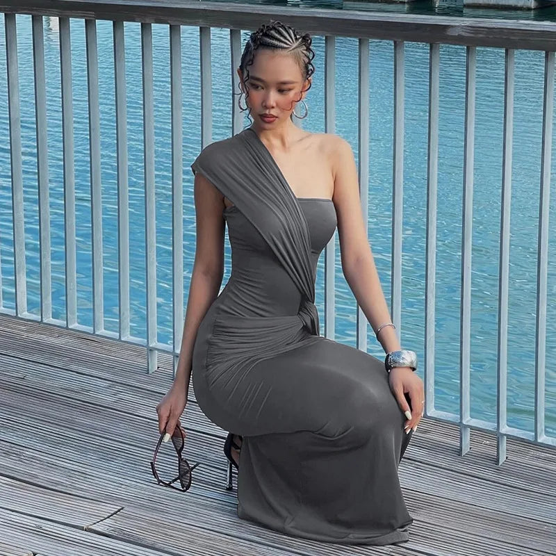 Woman in a gray one-shoulder dress sitting on a wooden deck with a blue water background.