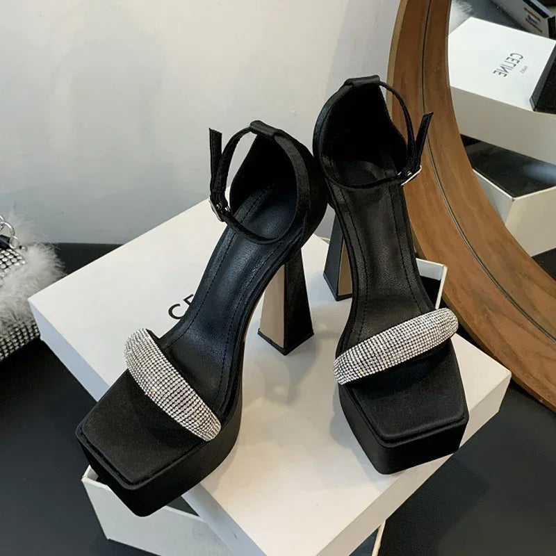 Black high-heeled sandals with decorative straps on a white platform, with a mirror and other items in the background.