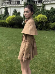 Woman in a beige outfit standing on grass with a building and greenery in the background