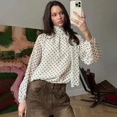 Woman taking a mirror selfie wearing a polka dot blouse and brown pants in a room.