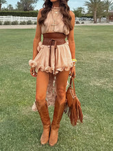 Woman in a beige top and skirt with brown boots and fringed bag standing on grass.