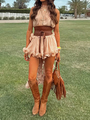 Woman in a beige top and skirt with brown boots and fringed bag standing on grass.