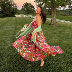 Woman in a floral dress standing in a grassy field with trees and a sunset in the background