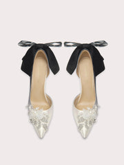 Pair of high-heeled shoes with black bows and pearl embellishments on a light gray background