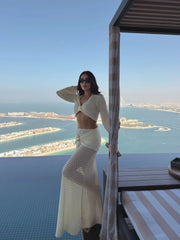 Woman in a white knit cropped long sleeve top and matching knit maxi skirt with draped detail, overlooking an ocean cityscape.