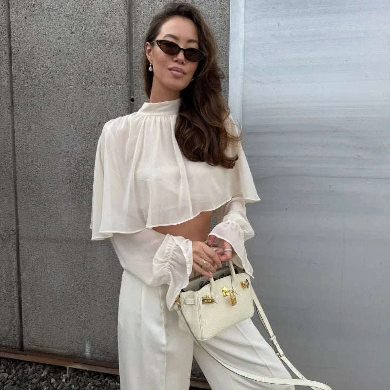 Woman in a white outfit holding a matching handbag against a gray wall.