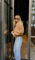 A woman wearing a khaki jacket and light blue jeans is standing in front of a glass door.