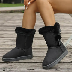 Black winter boots with fur trim worn by a person on a wooden floor.