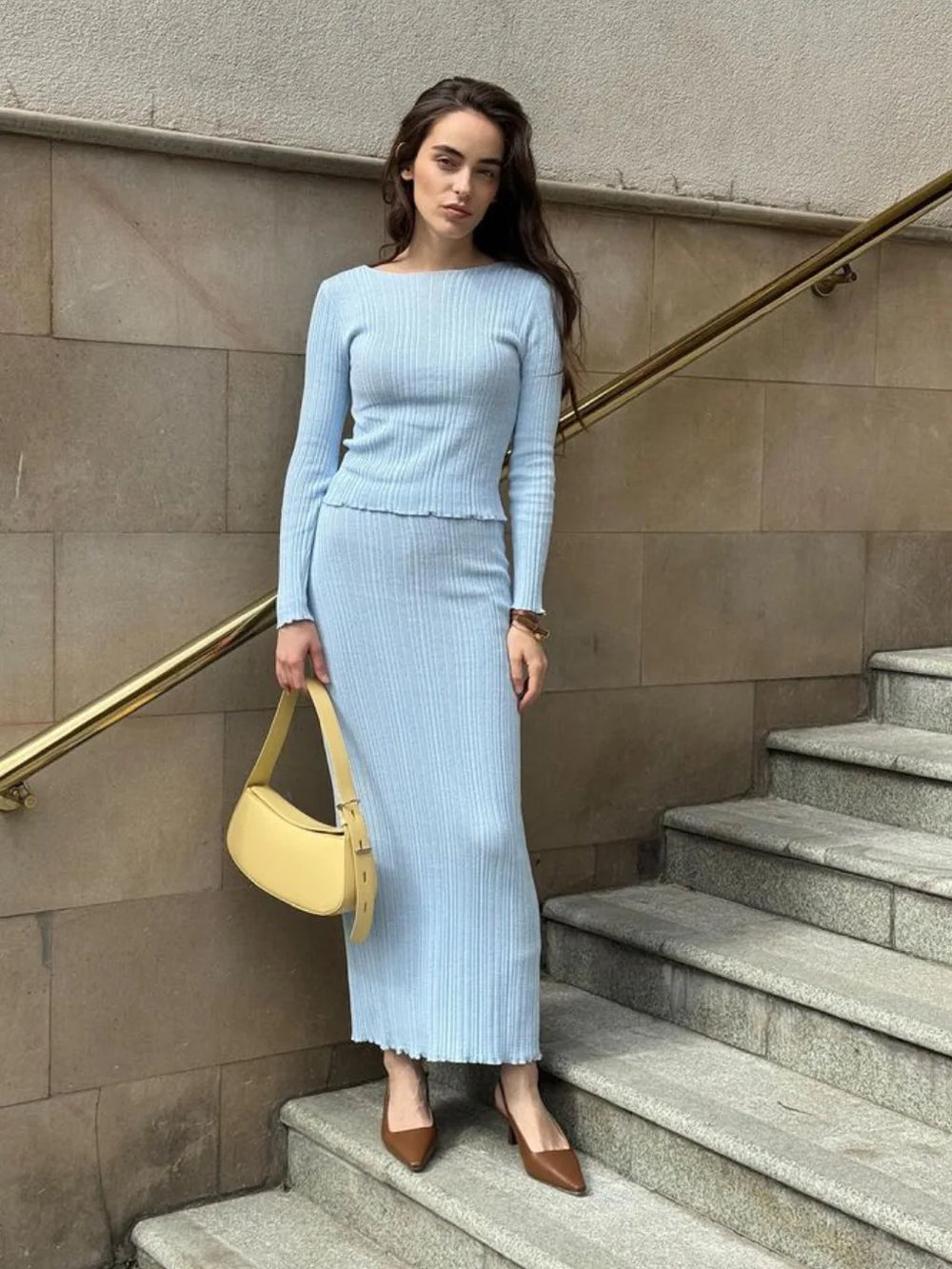 Woman in a light blue ribbed sweater and skirt standing on stone steps with a yellow handbag.