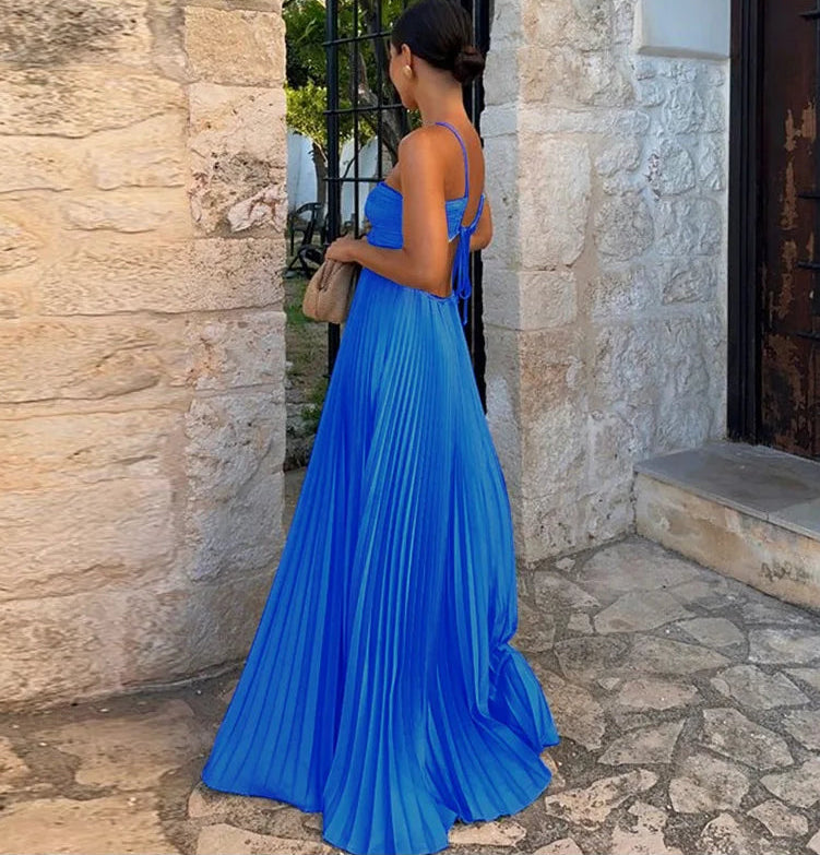 Woman in a blue pleated dress standing against a stone wall.