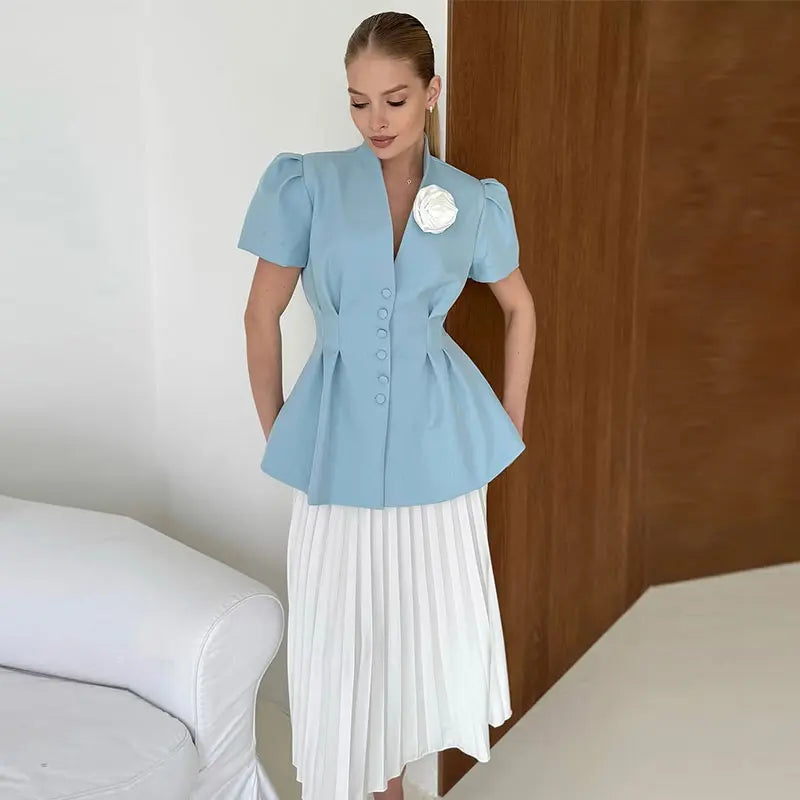 Woman wearing a light blue top with puff sleeves and a white pleated skirt, standing against a neutral background.