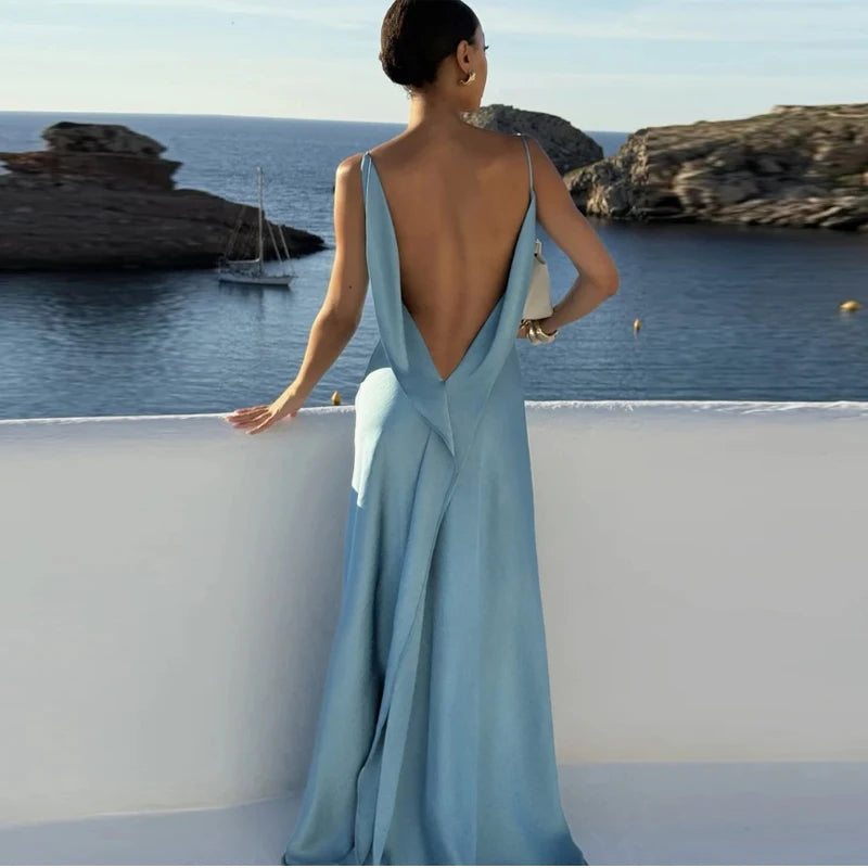 Woman in a light blue evening gown with an open back, standing by a scenic waterfront.