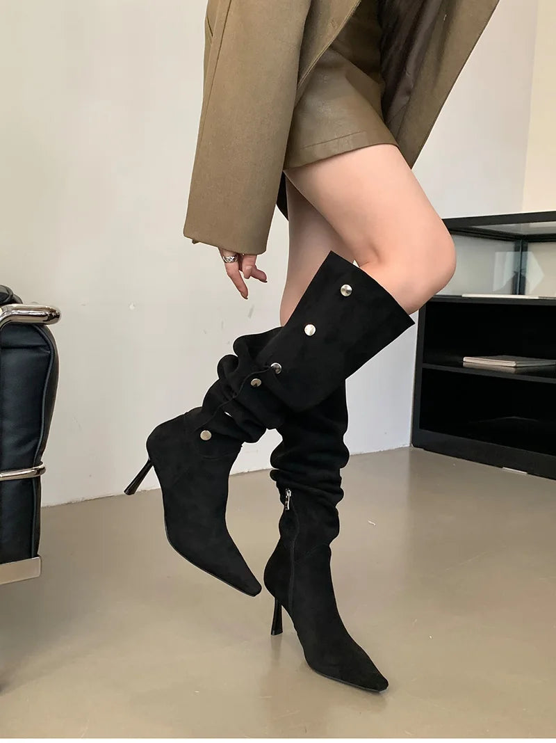 Person wearing black knee-high boots with high heels in a minimal indoor setting.