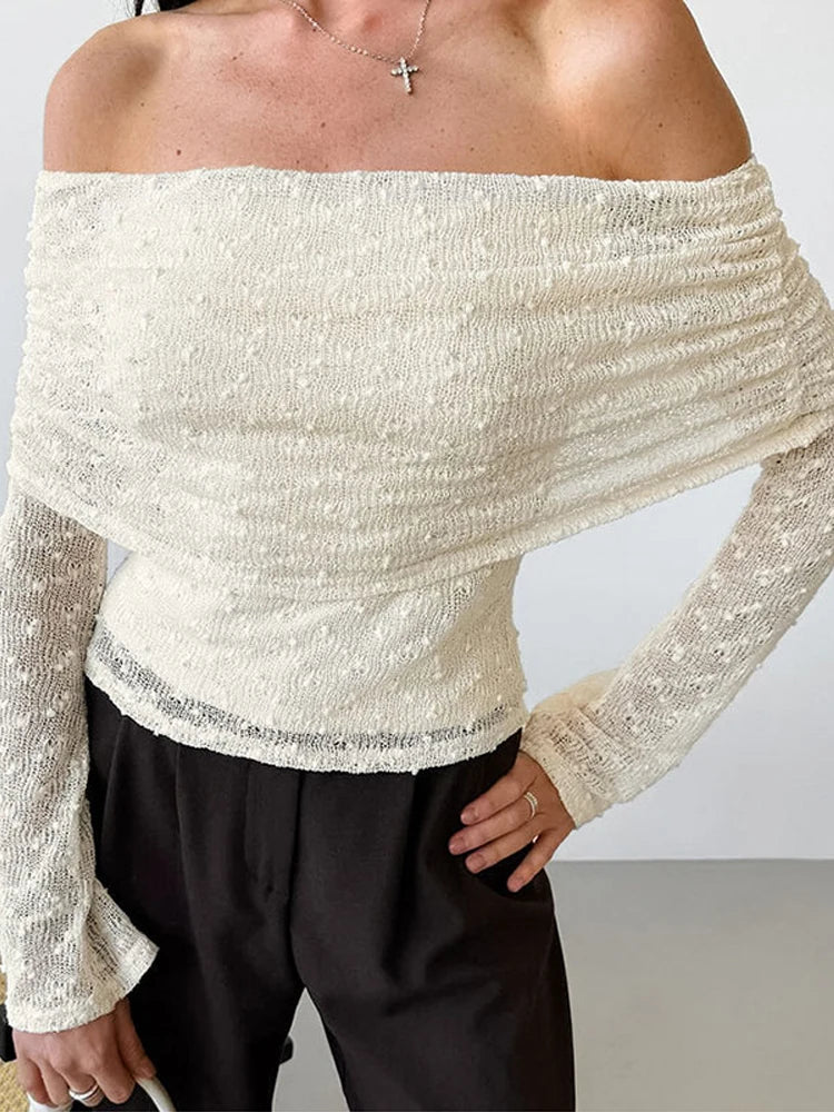 Person wearing a textured off-shoulder top with long sleeves and black pants against a plain background