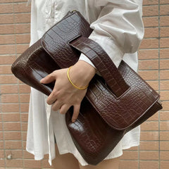 Brown textured clutch bag held by a person against a brick wall.