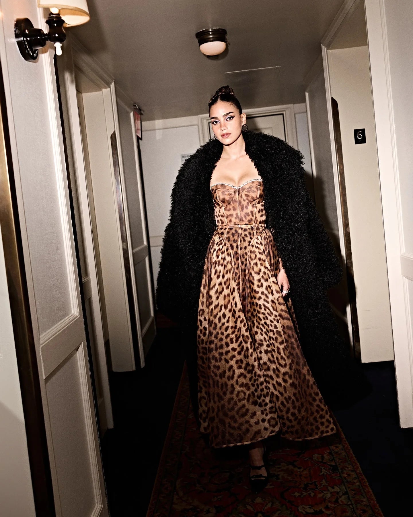 Woman wearing a leopard print dress and black coat in a hallway.