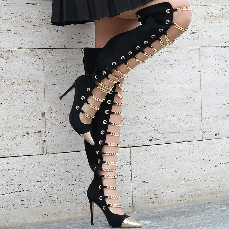 Black high-heeled boots with gold chain details worn by a person against a light stone wall.