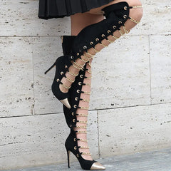 Black high-heeled boots with gold chain details worn by a person against a light stone wall.