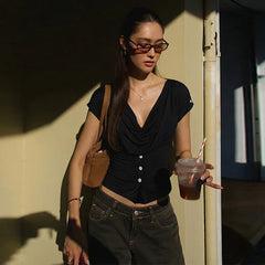 Woman in black top and jeans holding a drink, standing against a wall.