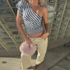 Person wearing a striped one-shoulder top and light pants, holding a pink fluffy bag against a glass block wall.