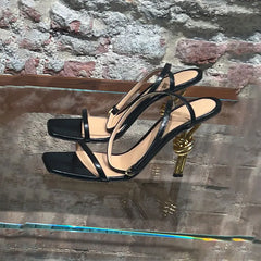 Black high-heeled sandals with gold heel on a reflective surface with a stone wall background