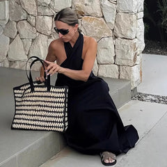 Woman in a black dress holding a black and white woven bag against a stone wall.