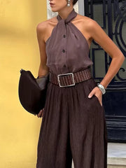 Woman wearing a brown halter top and wide-leg pants with a studded belt, holding a black handbag against a yellow wall.