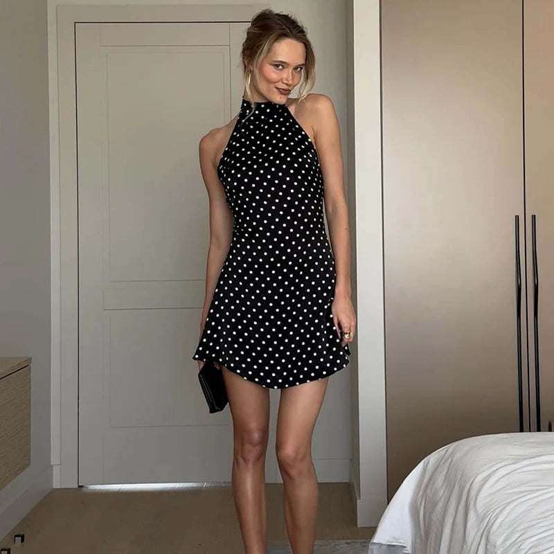 Woman wearing a black polka dot dress standing in a room with neutral walls and a bed.