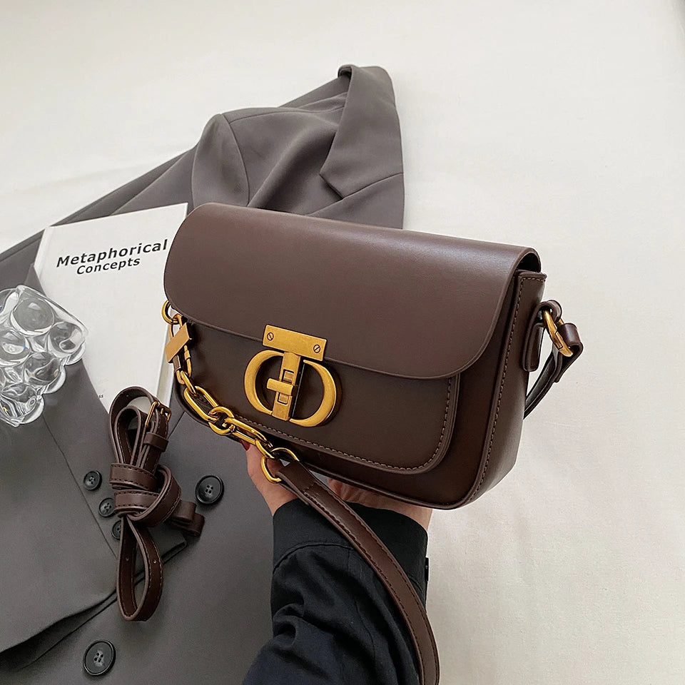 Brown leather handbag with gold chain held by a person, on a light gray background.