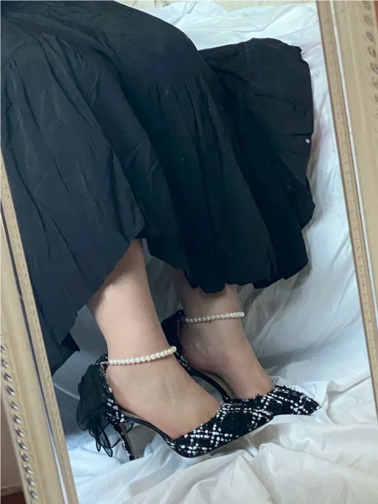 Person wearing black high-heeled shoes with bow details and pearl anklets, sitting on a white surface.