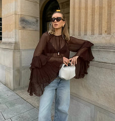 Woman in a brown ruffled blouse and light blue jeans holding a white handbag, standing against a stone building.