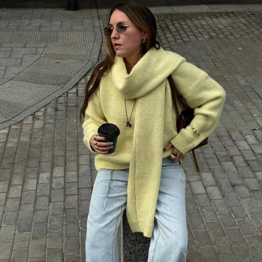 Woman in a yellow sweater and light blue jeans walking on a sidewalk, holding a black coffee cup.